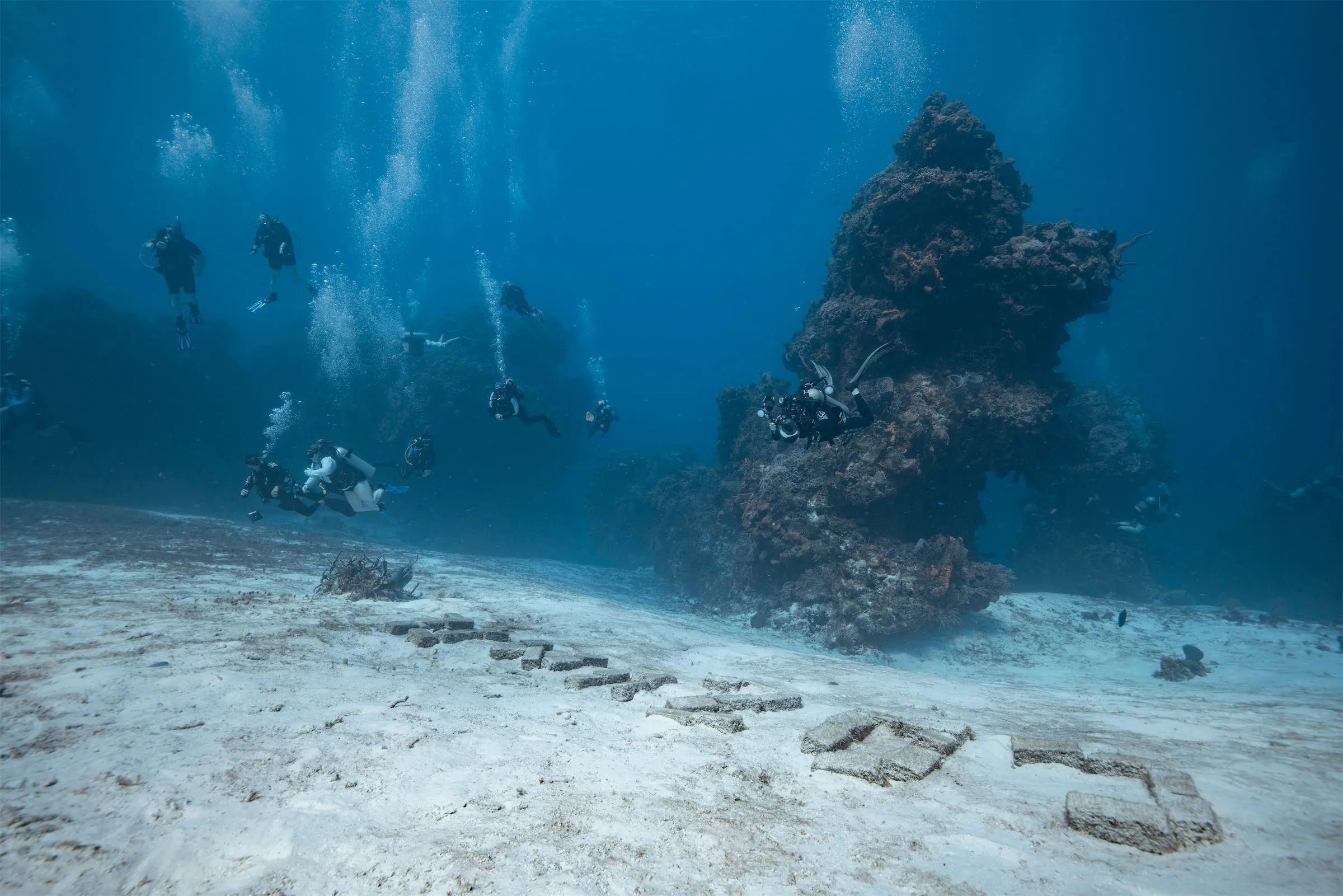 Rectangular bricks lying on white sand at Palancar Bricks, Cozumel, arranged into letters, with a coral pinnacle and a group of divers drifting past above